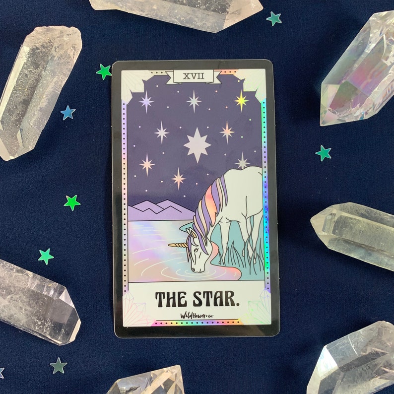May include: A tarot card with a holographic finish depicting a white unicorn drinking from a blue lake under a starry night sky. The card is labeled "The Star" and has the Roman numeral XVII in the top right corner.