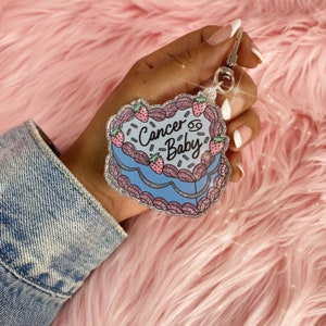 May include: A blue and pink glitter keychain shaped like a cake with the text "Cancer Baby" and a Cancer zodiac symbol.