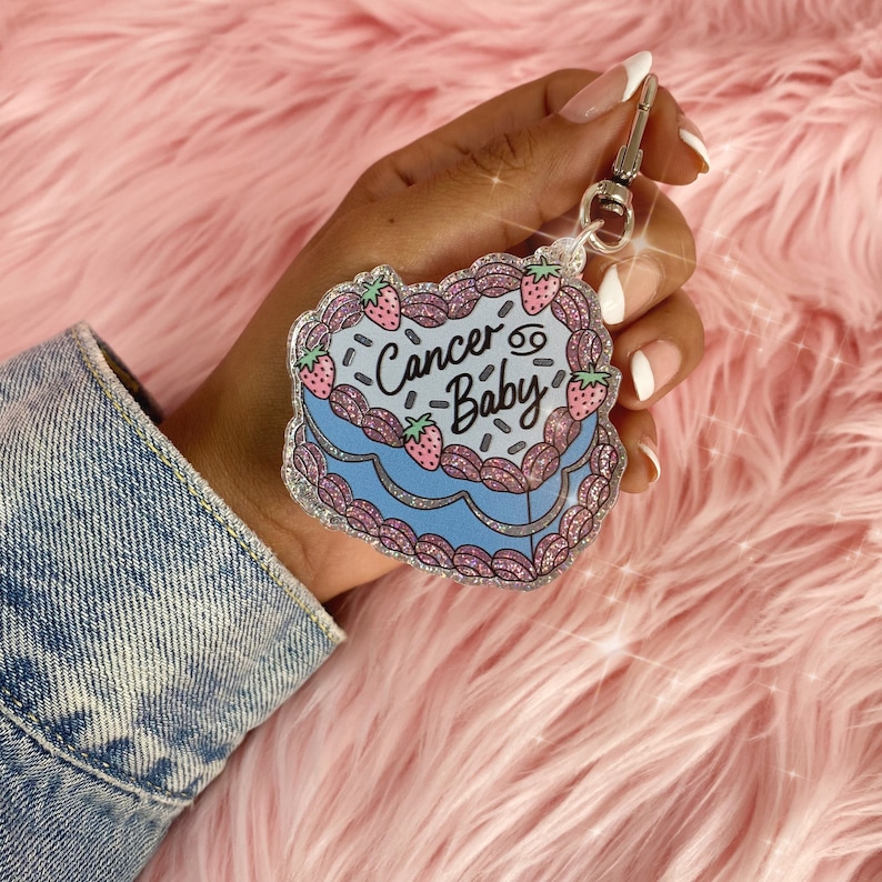 May include: A blue and pink glitter keychain shaped like a cake with the text "Cancer Baby" and a Cancer zodiac symbol.
