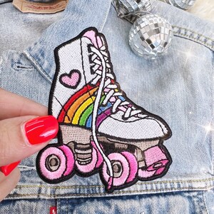 May include: Embroidered patch of a white roller skate with a rainbow and a pink heart. The wheels and laces are pink. The patch is on a denim jacket.