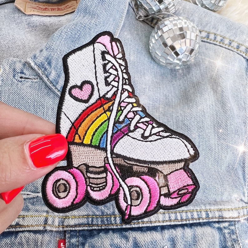 May include: Embroidered patch of a white roller skate with a rainbow and a pink heart. The wheels and laces are pink. The patch is on a denim jacket.
