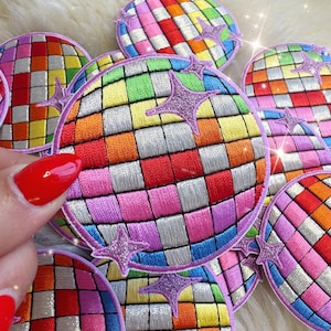 Peut inclure: Écussons brodés représentant une boule disco. Les écussons ronds présentent un motif quadrillé de différentes couleurs, notamment le rouge, l'orange, le jaune, le vert, le bleu et le violet. Chaque écusson a une bordure lavande et des étoiles lavande.