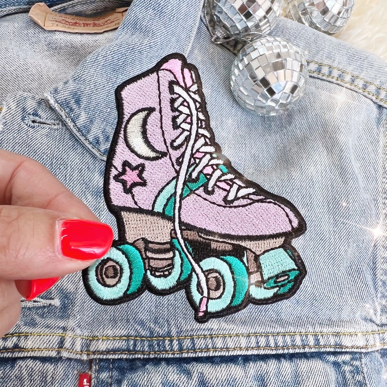May include: Embroidered patch of a pink roller skate with teal wheels and accents. The skate features a crescent moon and star design. White laces and a light blue denim jacket are in the background.