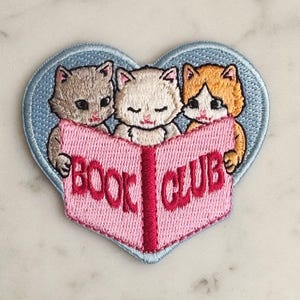 May include: Embroidered patch in the shape of a heart with three kittens reading a book. The book is pink with the words "BOOK CLUB" in red. The heart is outlined in light blue.