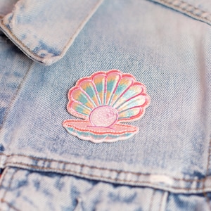May include: An embroidered patch depicting an open seashell with a pearl, in shades of pink, blue, and yellow. The patch is attached to a light blue denim jacket.
