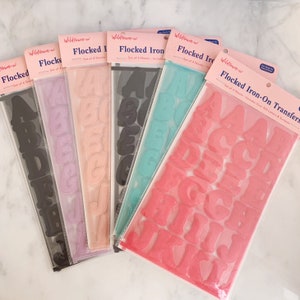 May include: Six packages of Wildflower + Co. flocked iron-on transfers, each containing four sheets of letters and symbols. The sets feature various colors including black, lavender, peach, dark gray, teal, and pink, all arranged in clear plastic sleeves.