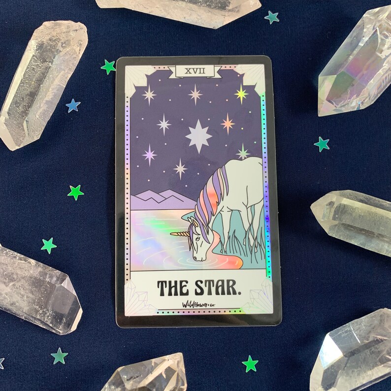 May include: A holographic tarot card with a black border and a purple and white unicorn drinking from a lake. The card is titled "The Star" and has the Roman numeral XVII in the top right corner. The background is a dark blue sky with white stars.