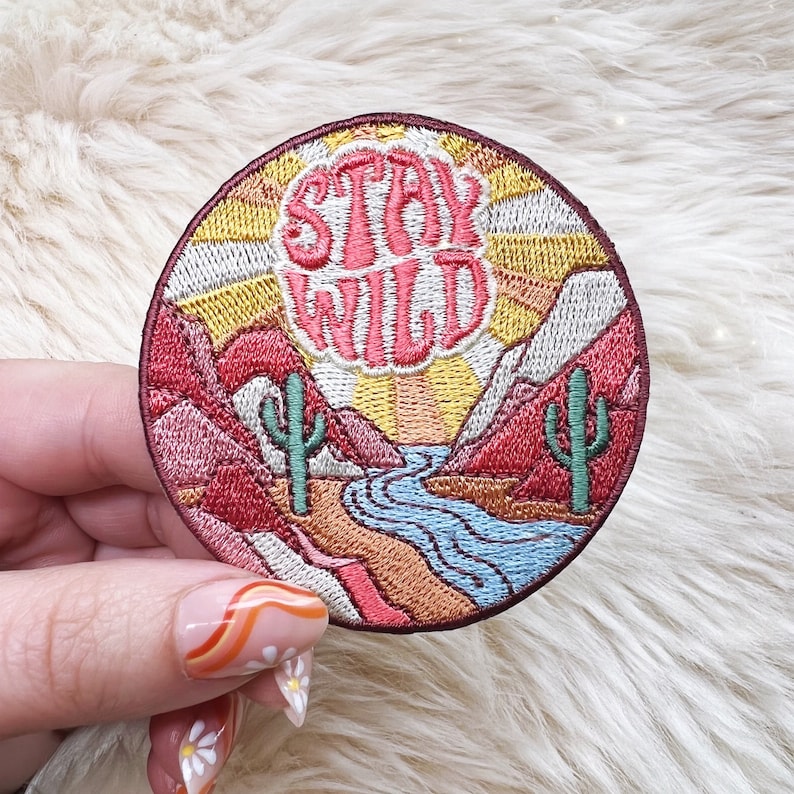Peut inclure: &Eacute;cusson brod&eacute; avec un motif de paysage d&eacute;sertique. L'&eacute;cusson repr&eacute;sente une rivi&egrave;re, des montagnes, des cactus et les mots "Stay Wild" dans une police r&eacute;tro. La palette de couleurs comprend des nuances de rouge, d'orange, de jaune, de vert et de bleu.