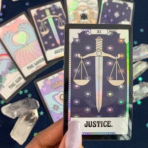 May include: A holographic tarot card with a black border and a purple background. The card depicts a scale with a sword, surrounded by eyes. The card number "XI" and the word "JUSTICE" are printed on the card.