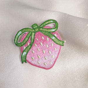 May include: A pink embroidered strawberry with a green bow. The strawberry has white seeds and a green stem.