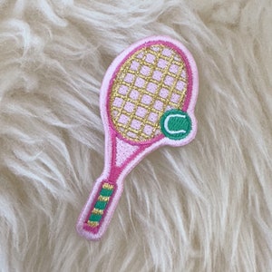 Peut inclure: Patch brodé rose et or représentant une raquette de tennis avec une balle de tennis verte sur laquelle est inscrit un "C".