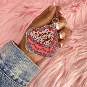 May include: Pink glitter cake keychain with the text "Scorpio Baby" and a strawberry design.