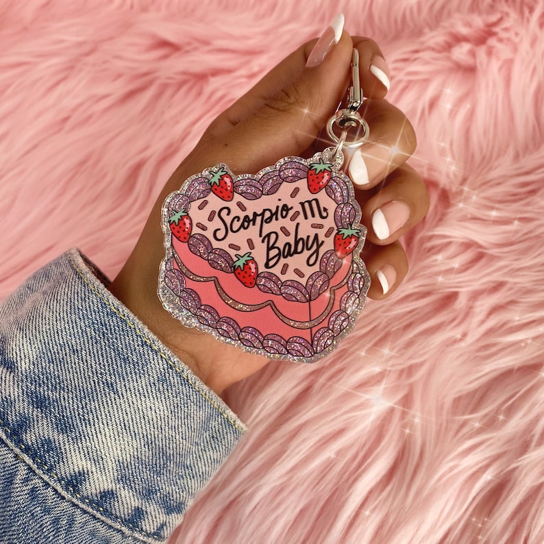 May include: Pink glitter cake keychain with the text "Scorpio Baby" and a strawberry design.