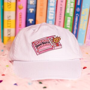 Puede incluir: Gorra de b&eacute;isbol blanca con un dise&ntilde;o bordado de una caja de cerillas rosa con el texto "Slow Burn" y una cerilla encendida. La gorra se encuentra frente a un fondo de libros coloridos y una superficie rosa y esponjosa.