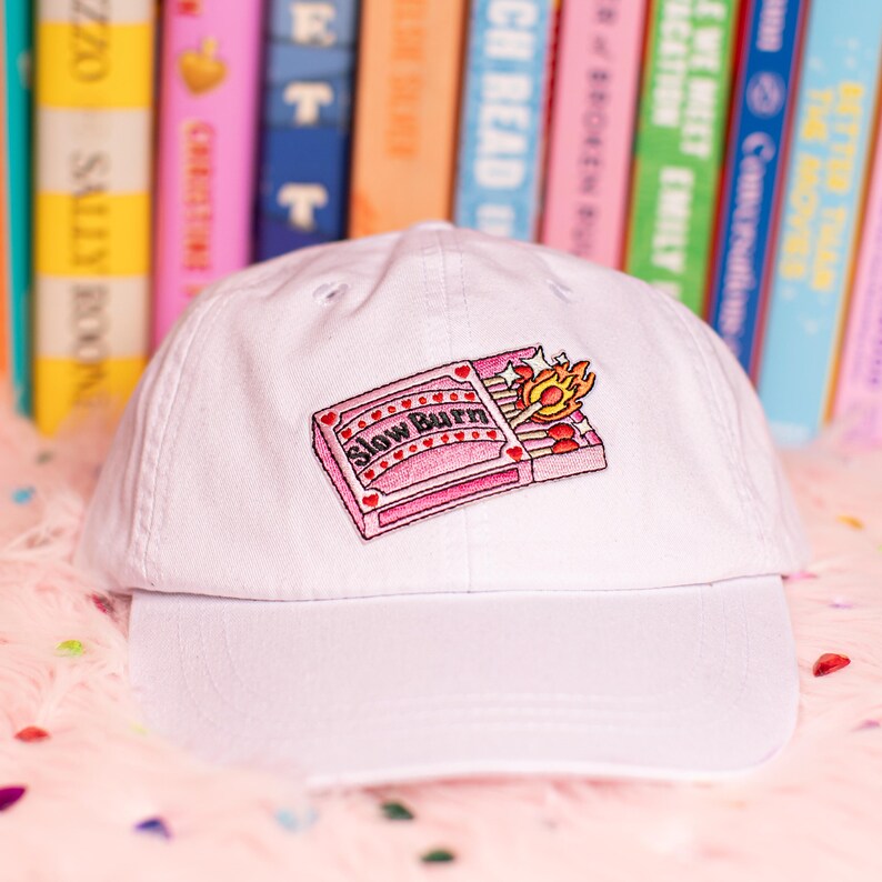Puede incluir: Gorra de b&eacute;isbol blanca con un dise&ntilde;o bordado de una caja de cerillas rosa con el texto "Slow Burn" y una cerilla encendida. La gorra se encuentra frente a un fondo de libros coloridos y una superficie rosa y esponjosa.