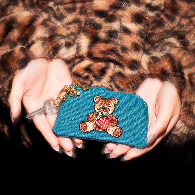 May include: Teal key pouch with a gold zipper and a brown teddy bear holding a red strawberry. A key is attached to the pouch. The background is a brown and black leopard print.