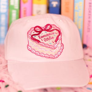 May include: A pale pink baseball cap with a cake-shaped embroidered patch. The patch has a pink bow and the words "Romance Girlie". The cap is made of a soft material and is suitable for casual wear.