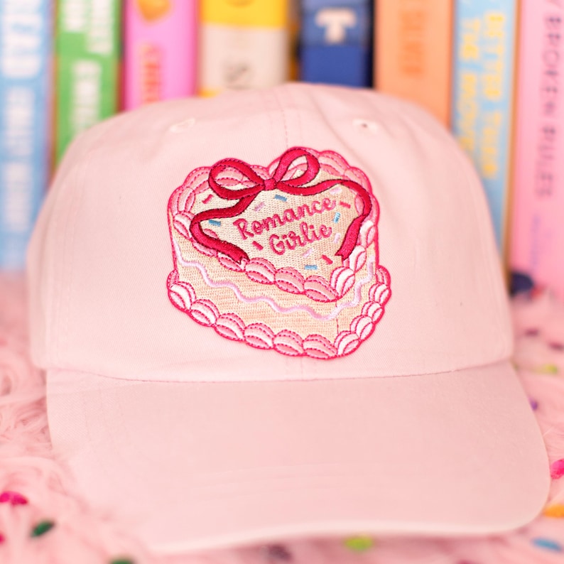May include: A pale pink baseball cap with a cake-shaped embroidered patch. The patch has a pink bow and the words "Romance Girlie". The cap is made of a soft material and is suitable for casual wear.