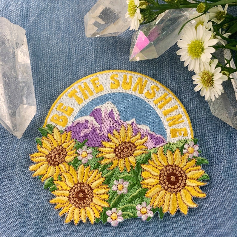 May include: Embroidered iron-on patch with a yellow, white, and green design of sunflowers and a mountain range. The text "Be the Sunshine" is in a white circle around the design.