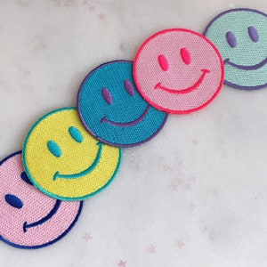 Puede incluir: Cinco parches bordados con caritas sonrientes en diferentes colores. Los parches son amarillos, rosas, azules, rosas y verdes. Cada parche tiene un tono de morado diferente para los ojos y la boca.