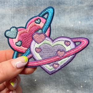 May include: Two embroidered iron-on patches in the shape of hearts with a ring around them, representing planets. The hearts are pink, purple, and blue with glitter accents.