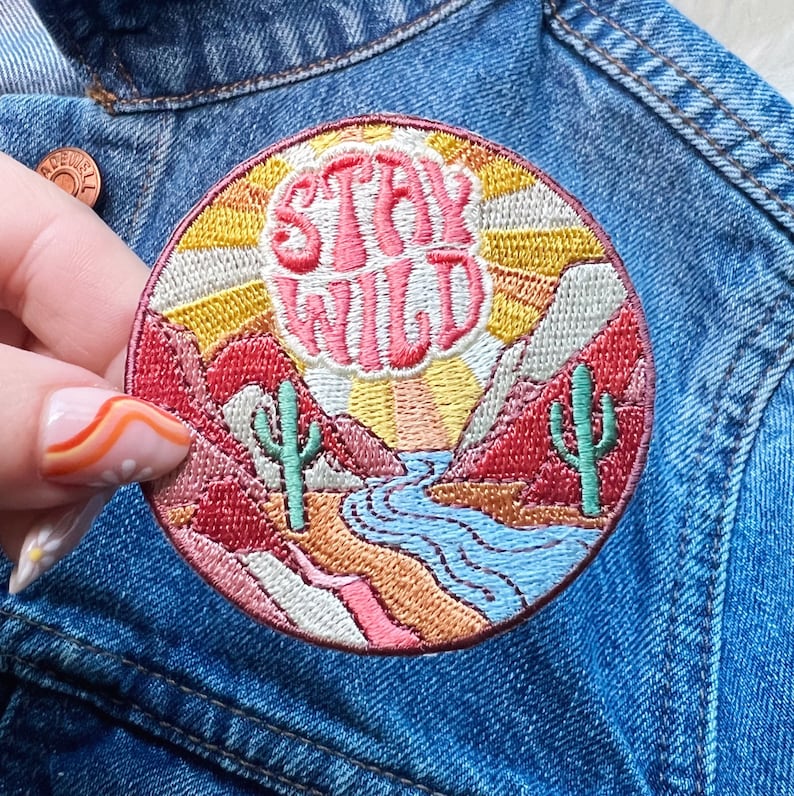 Peut inclure: &Eacute;cusson rond brod&eacute; avec les mots "STAY WILD" en script rose. Le patch repr&eacute;sente un paysage d&eacute;sertique avec des montagnes, des cactus, une rivi&egrave;re et un coucher de soleil dans des tons de rouge, jaune et bleu. Le patch est cousu sur une veste en jean.