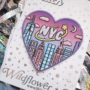 May include: A heart-shaped iron-on patch featuring a colorful New York City skyline illustration with the text NYC in white letters surrounded by a yellow ring and star. The patch is mounted on a white card labeled Iron-On Patch Wildflower Co.