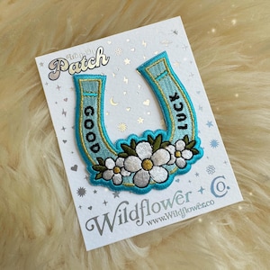 May include: A blue horseshoe shaped iron-on patch with white flowers and the words "Good Luck" in black lettering. The patch is on a white card with the words "Iron-On Patch" and "Wildflower Co." in gold lettering.