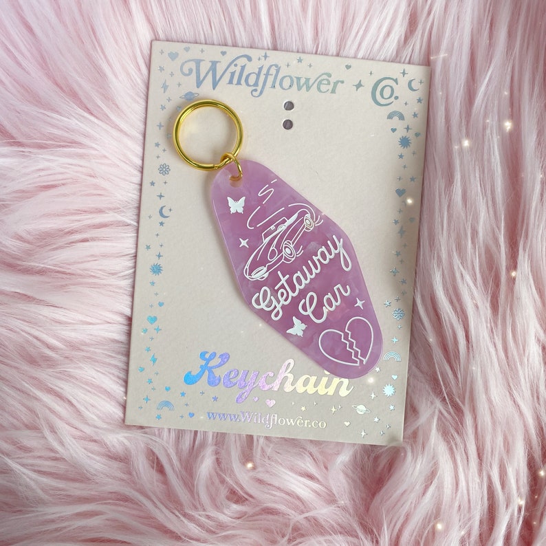 May include: A lavender-colored keychain with a gold ring, featuring a car graphic and the words "Getaway Car." The keychain also includes butterfly and broken heart designs. The card behind the keychain says "Wildflower Co." and "Keychain."