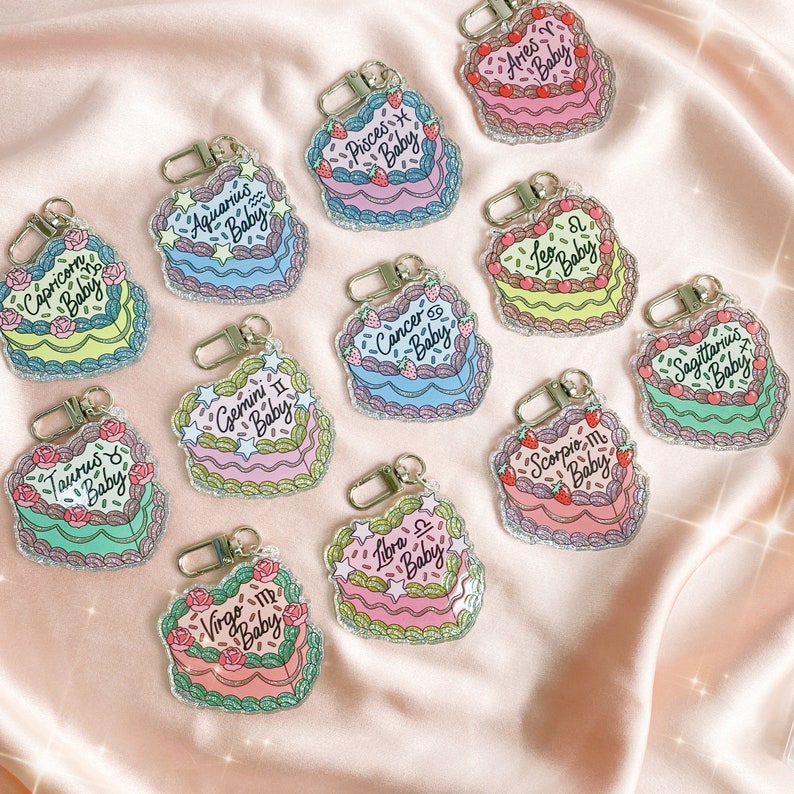 May include: Twelve heart-shaped keychains with glitter and a cake design. Each keychain features a different zodiac sign with the text "Baby" underneath. The keychains are arranged in a circle on a pink background.