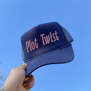 May include: A navy blue trucker hat with a pink embroidered design that says "Plot Twist".