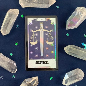 May include: A tarot card with a holographic finish depicting the Justice card. The card is dark blue with a gold sword and scales. The card number, XI, is in the top right corner. The text "JUSTICE." is printed in white at the bottom of the card.