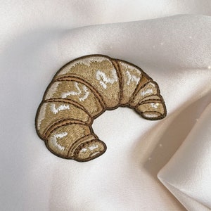Peut inclure: Un patch brodé marron en forme de croissant. Le croissant est bordé de fil marron et présente des détails de couture blancs.