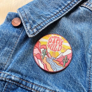 Peut inclure: Un &eacute;cusson rond brod&eacute; avec l'inscription "STAY WILD". Le patch repr&eacute;sente un paysage d&eacute;sertique avec des cactus, des montagnes et une rivi&egrave;re, sur fond de coucher de soleil. Il est fix&eacute; sur une veste en jean bleue.