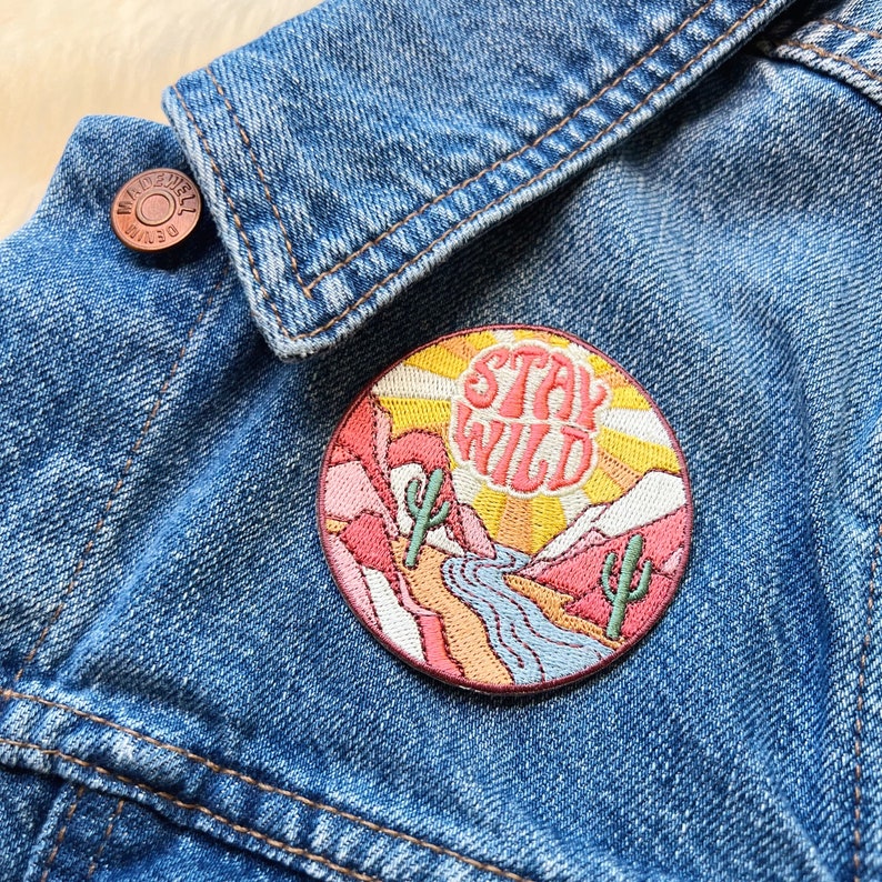 Peut inclure: Un &eacute;cusson rond brod&eacute; avec l'inscription "STAY WILD". Le patch repr&eacute;sente un paysage d&eacute;sertique avec des cactus, des montagnes et une rivi&egrave;re, sur fond de coucher de soleil. Il est fix&eacute; sur une veste en jean bleue.