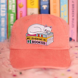 May include: A coral baseball cap with an embroidered design. The design features a white cat sleeping on a stack of books. The text "MY EVENING IS BOOKED" is below the cat. The cap has a curved peak.