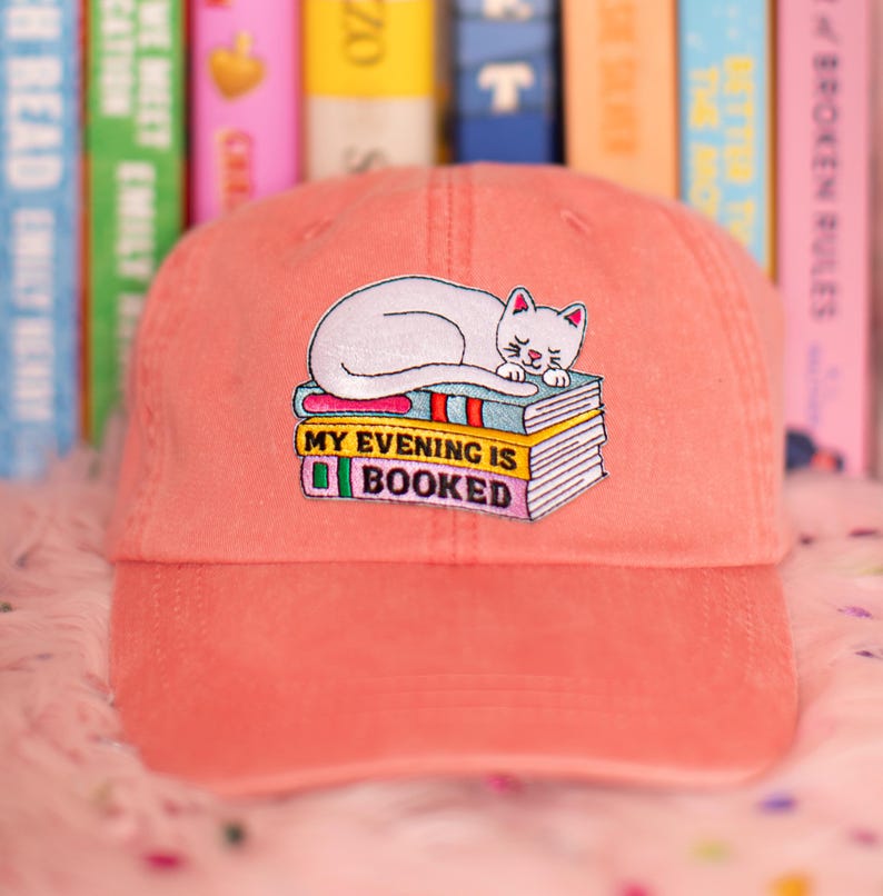 May include: A coral baseball cap with an embroidered design. The design features a white cat sleeping on a stack of books. The text "MY EVENING IS BOOKED" is below the cat. The cap has a curved peak.