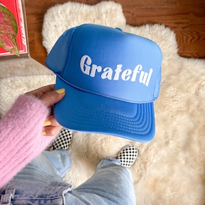 May include: A blue trucker hat with a white mesh back and the word "Grateful" in white retro-style lettering.