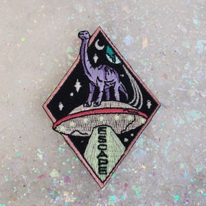 May include: An embroidered patch featuring a purple dinosaur standing on a flying saucer with a green planet and a crescent moon in the background. The patch is in the shape of a diamond and has a pink outline.