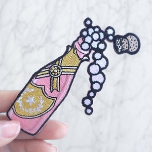 Peut inclure: Bouteille de champagne rose avec des accents dorés et un bouchon blanc et noir qui éclate avec des bulles. La bouteille est brodée d'une étoile dorée et d'un ruban doré.