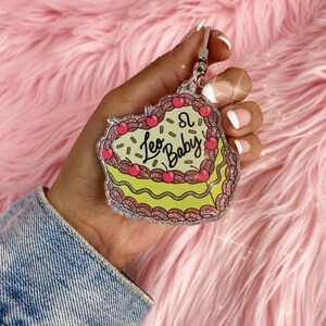 May include: A glittery pink and yellow heart-shaped keychain with the text "Leo Baby" and the astrological symbol for Leo. The keychain is made of clear acrylic and has a silver key ring.