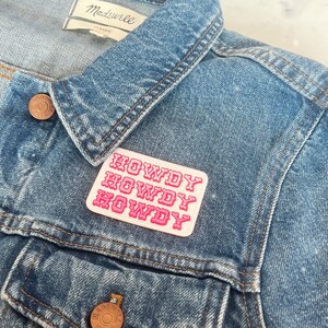 May include: A pink and white embroidered patch with the word "Howdy" repeated three times, sewn onto a blue denim jacket.