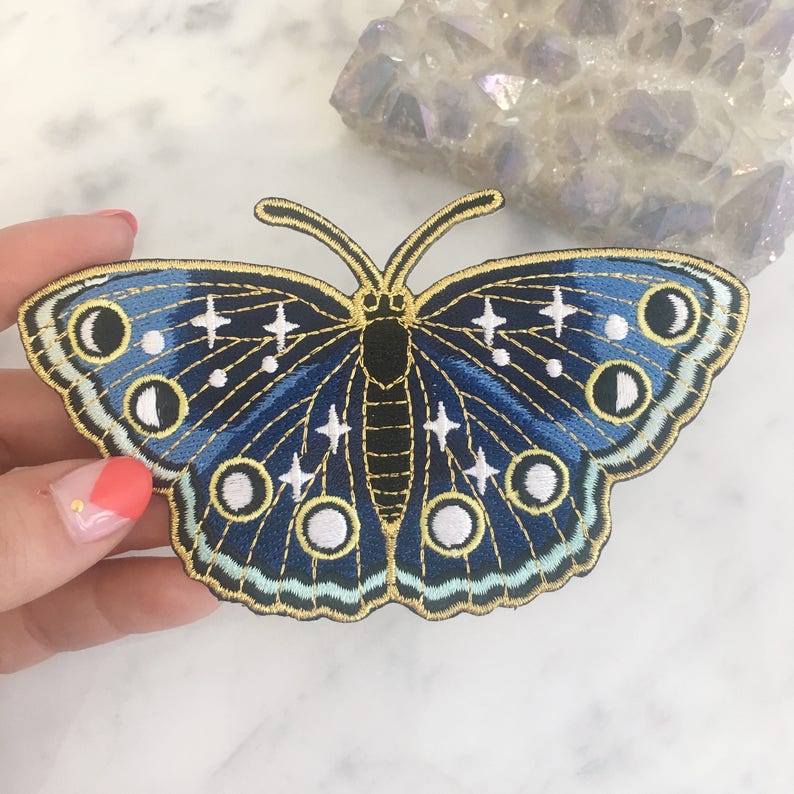 以下が含まれることがあります： 月と星が刺繍された青と金の蝶のワッペン。蝶は金糸で縁取られており、黒い体をしている。