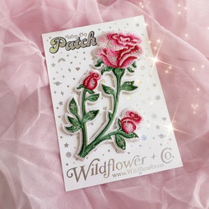May include: Iron-on patch featuring a pink rose with green leaves and a white stem. The patch is labeled "Iron-on Patch" and "Wildflower Co." with the website address "www.wildflower.co".