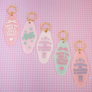 Pode incluir: Cinco chaveiros retrô em rosa, branco e verde menta, com argolas douradas. Cada chaveiro tem uma frase diferente em cores pastel, incluindo "OMW TO GET A LITTLE TREAT" e "RUNNING ON CAFFEINE & CHAOS". Fundo rosa quadriculado.