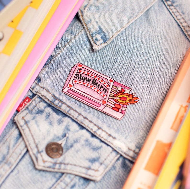 May include: A pink embroidered patch with the words "Slow Burn" on a denim jacket. The patch shows a matchbook with lit matches and flames. Other colourful items are in the background.
