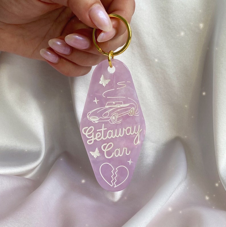 May include: A lavender motel-style keychain with a gold ring. The keychain features a white car graphic, butterflies, stars, and the words "Getaway Car." A broken heart graphic is at the bottom.