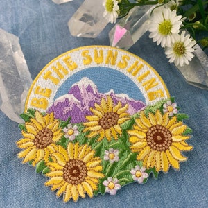 May include: Embroidered patch with a yellow, green, and purple mountain scene and the text "Be the Sunshine". The patch features three yellow sunflowers with brown centers.