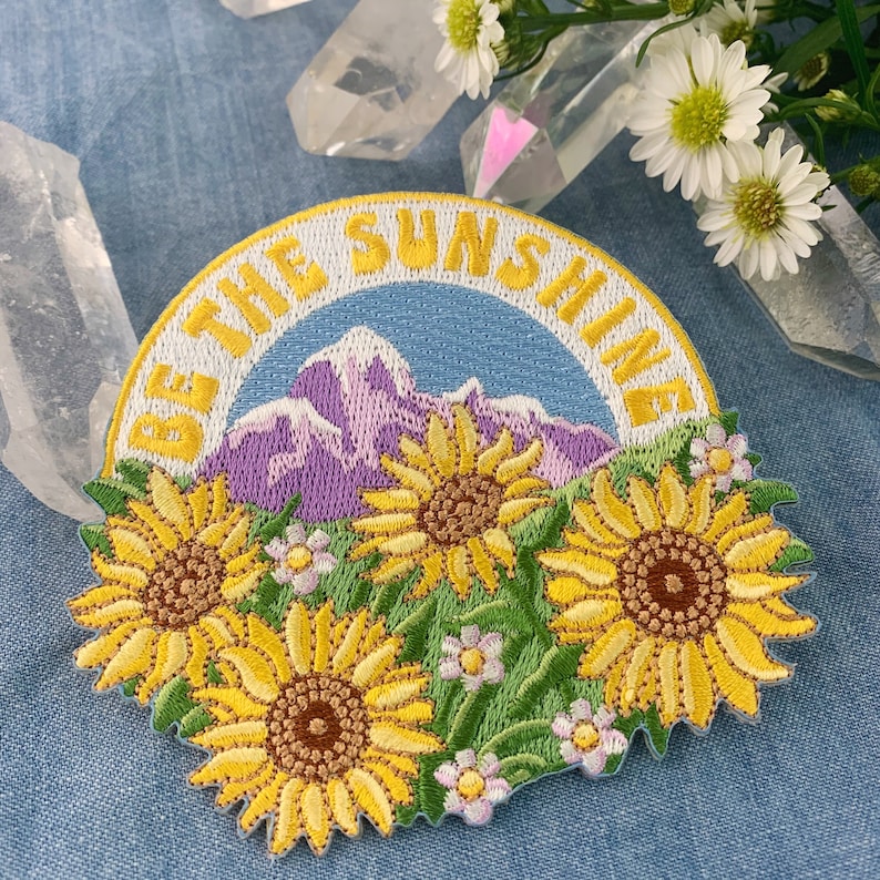May include: Embroidered patch with a yellow, green, and purple mountain scene and the text "Be the Sunshine". The patch features three yellow sunflowers with brown centers.