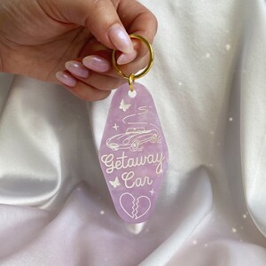 May include: A lavender-colored keychain with a gold ring. The keychain features a car graphic, butterflies, stars, and the words "Getaway Car" in white script. A broken heart graphic is at the bottom.